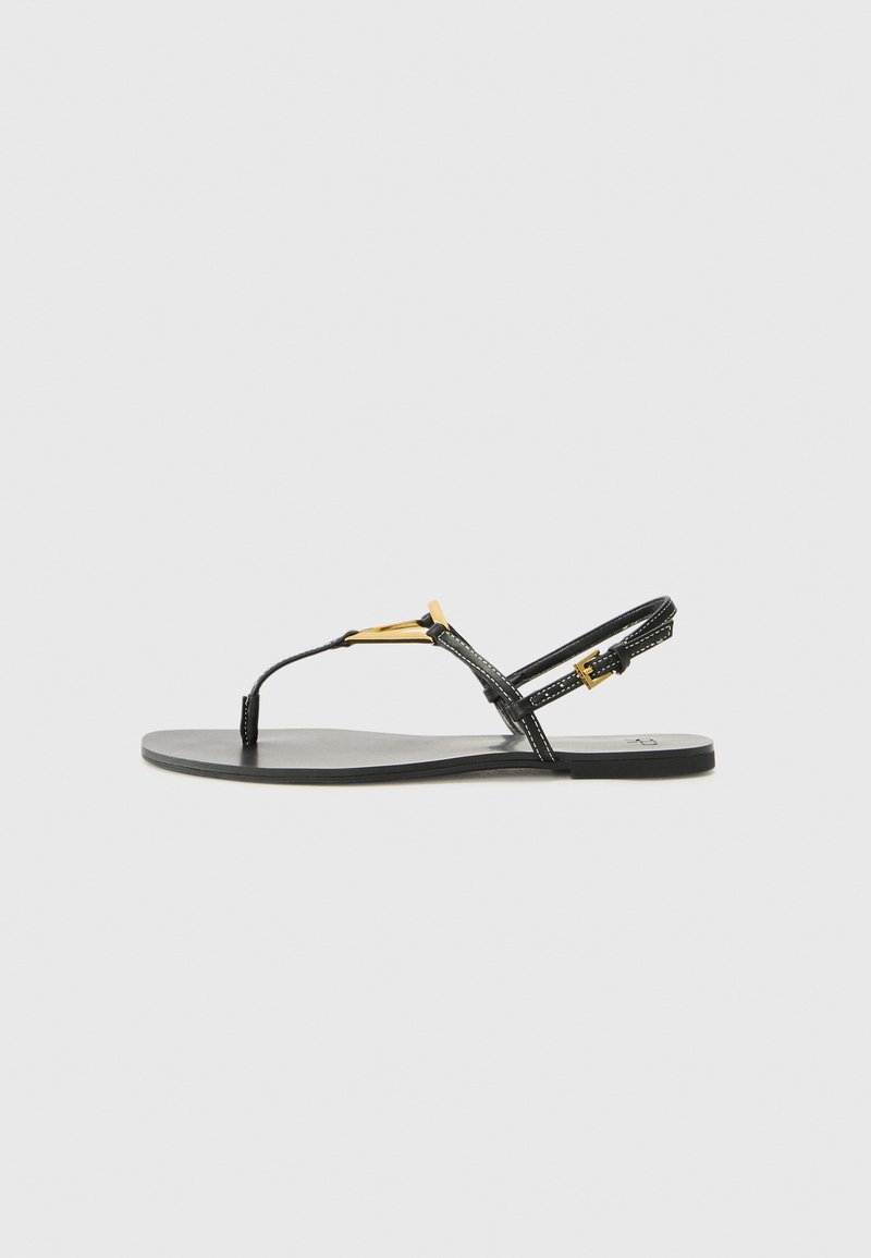 Sandali neri piatti con design a T-strap, caratterizzati da un accento dorato e una parte superiore testurizzata. La suola è liscia e minimalista.