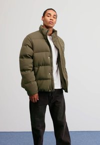 Giacca puffer verde oliva con texture a trapunte, bottoni a pressione, colletto alto, indossata su una maglietta bianca e pantaloni neri.
