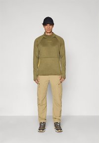 Dlhý rukáv olivovo zelený top s textúrovaným vzorom a žltými detailmi, v kombinácii s svetlo béžovými cargo nohavicami a tmavými športovými topánkami.