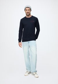 Granatowy t-shirt z długim rękawem z czerwonym logo, w połączeniu z jasnoniebieskimi jeansami i białymi sneakersami z czarnymi akcentami. Szary, prążkowany beanie.