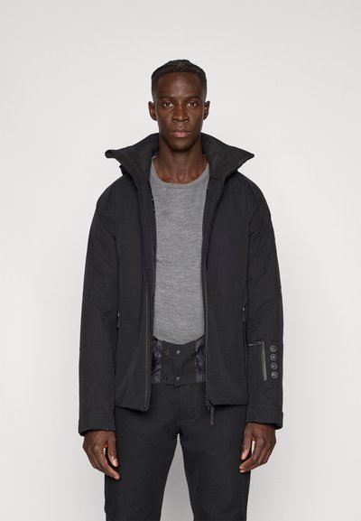 Čierna zateplená bunda s vysokým golierom, zapínaním na zips a bočnými vreckami. Nosená cez sivé tričko, s textúrovaným povrchom tkaniny.