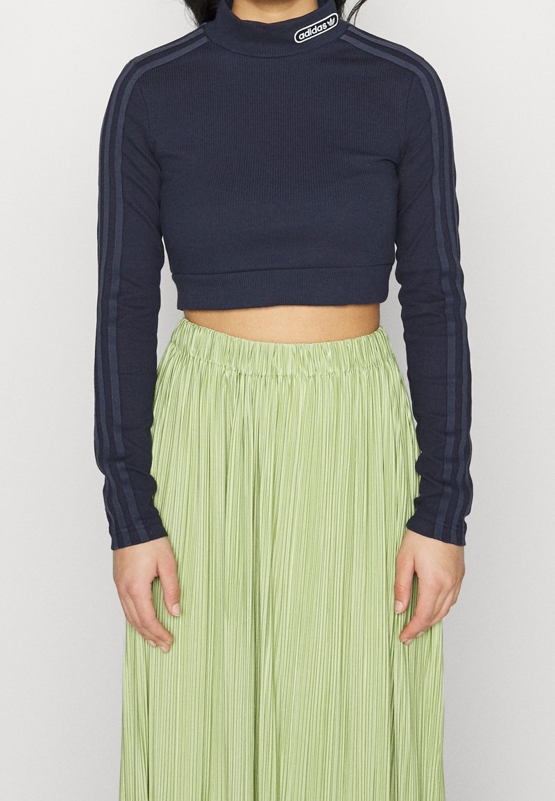 Marineblaues, geripptes Crop-Top mit langen Ärmeln und Stehkragen, das mit dunkelblauen Streifen und einem Adidas-Logo-Patch versehen ist, kombiniert mit einem plissierten grünen Rock.