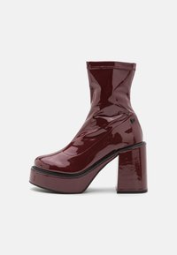 Botín de tobillo de charol burdeos con un alto tacón grueso, diseño elegante y costura en bruto. Presenta un acabado texturizado y cremallera lateral.