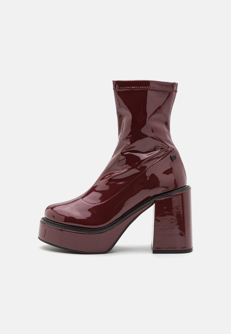 Botín de tobillo de charol burdeos con un alto tacón grueso, diseño elegante y costura en bruto. Presenta un acabado texturizado y cremallera lateral.