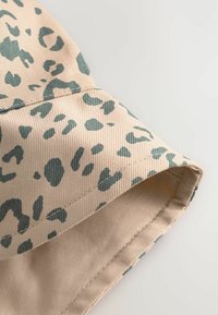 Chapeau seau tan en tissu texturé, orné d'un motif léopard vert. La bordure est légèrement incurvée, mettant en valeur le design.