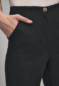 Zwarte op maat gemaakte broek met een gladde textuur, voorzien van een voorzak, opvallende verticale naden en een gouden knoopsluiting bij de taille.