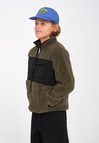 Veste en polaire de couleur vert olive avec un panneau noir sur la poitrine, fermeture éclair, deux poches avant et col haut ; portée avec une casquette bleue.
