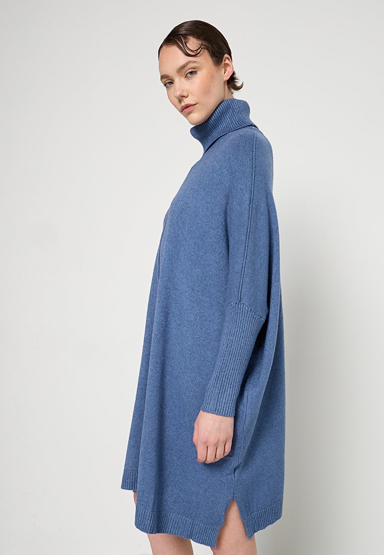 Frau mit kurzen Haaren, die ein lockeres blaues Rollkragenkleid trägt und seitlich vor einem schlichten Hintergrund steht.