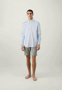 Jovem em pé descalço, vestindo uma camisa de botão azul clara e calções cinzentos até aos joelhos, contra um fundo simples.