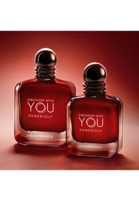 To mørkerøde parfymeflasker med runde sølvkorker, merket "Stronger With You Powerfully" av Emporio Armani på en rød gradientbakgrunn.