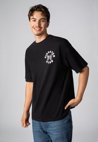 Schwarzes Baumwoll-T-Shirt mit kurzen Ärmeln, das auf der linken Brust ein weißes grafisches Design eines Schmetterlings und den Text "DISASTER CLUB" zeigt.