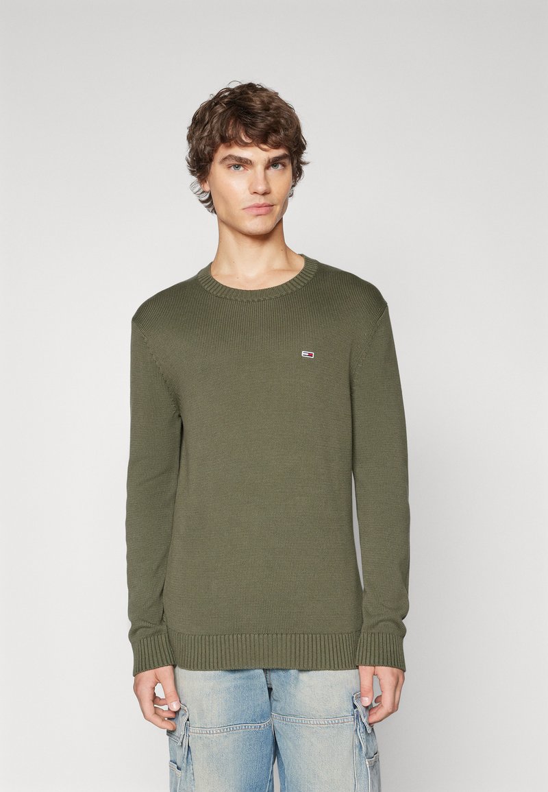 Pull en tricot côtelé vert olive avec un col rond et des manches longues, présentant un petit patch logo sur la poitrine. Tissu texturé et poignets ajustés.