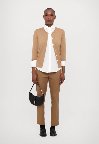 Beige cardigan over a white shirt, brown trousers, and black shoes. Black bag with a gold accent. Smooth texture, classic fit.