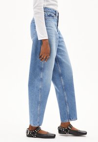 Personne portant un jean large taille haute bleu clair, un haut à manches longues blanc et des chaussures plates noires avec des brides à clous à la cheville.