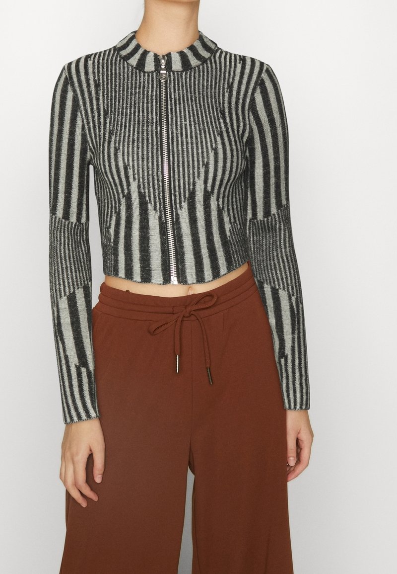 Cardigan cropped a righe nere e grigie con una texture a coste e colletto alto. Abbinato a pantaloni marroni a vita alta.