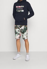 Mężczyzna w granatowej bluzie z kapturem Tommy Hilfiger z tropikalnym nadrukiem na logo oraz w dopasowanych szortach z tym samym tropikalnym wzorem, białe sneakersy i szare skarpetki.