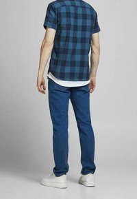 Man som bär en blå rutigt kortärmad skjorta, vit undertröja, blå jeans och vita sneakers, står vänd bort mot en grå bakgrund.