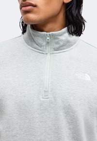 Personne portant une polaire gris clair avec une fermeture éclair sur un quart du devant et le logo The North Face sur la poitrine, fermé jusqu'au col.