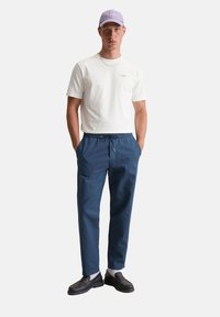 Casuele outfit bestaande uit een witte t-shirt, blauwe joggingbroek met trekkoord en zwarte instappers. Accessoires zijn een lichtpaarse pet en witte sokken.