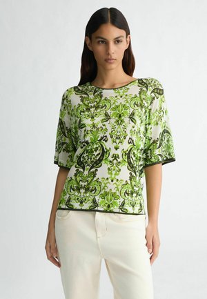 Korte mouwen groen en witte gebloemde top met een ronde hals, voorzien van ingewikkelde ontwerpen en contrasterende zwarte afwerking. Draagt een lichte broek.