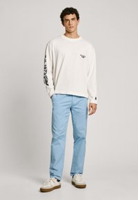 Balta krekla garajām piedurknēm ar melnu grafiku kreisajā piedurknē, apvienota ar gaišziliem biksēm un baltām kedām ar brūniem zoliem.