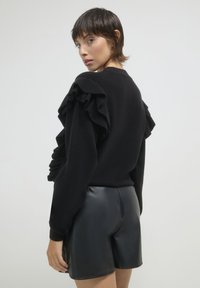 Pull en polaire noir avec détails à volants aux épaules, associé à un short en cuir noir taille haute, présenté sur un fond clair.