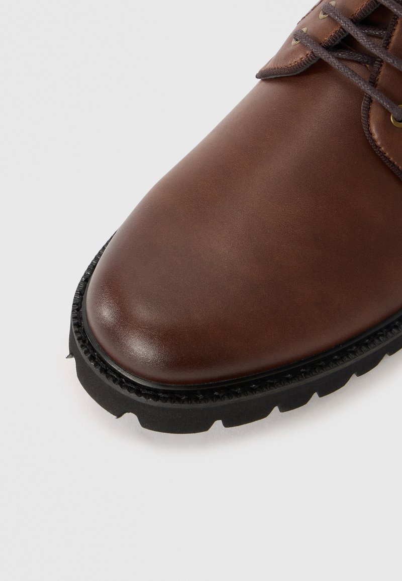 Zapato de cuero marrón con un acabado suave, puntera redondeada y suela de goma negra contrastante con detalles texturizados y borde cosido.