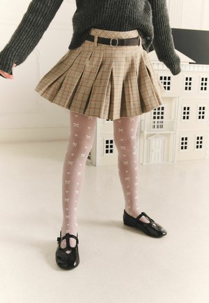 Kind draagt een bruine geplooide rok, geribbelde grijze trui, roze panty's met strikpatronen en zwarte Mary Jane schoenen, staand op een witte vloer.