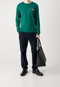 Zielona koszula z długim rękawem z logo, granatowe spodnie i czarne buty. Czarna torba na ramię trzymana w ręce, minimalistyczny design i gładka faktura.