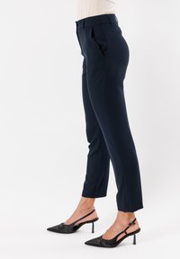 Pantaloni navy su misura con pinces, lunghezza cropped e texture liscia. Indossati con décolleté aperti neri a punta. Design minimalista.