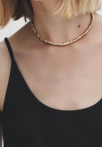 Collana choker in oro con un design ritorto, posizionata su una canotta nera a coste, che presenta una superficie liscia e una finitura lucida.