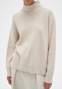 Beige turtleneck sweater made of soft fabric with a relaxed fit, featuring dropped shoulders and ribbed cuffs, paired with light-colored trousers.