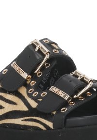 Infradito a strisce zebra nere e marroni con hardware dorato, fibbie laterali regolabili e suola spessa. Parte superiore testurizzata e dettagli in pelle.