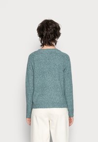 Pull tricoté couleur sarcelle avec un col rond et des manches longues. Présente une finition texturée et un ourlet côtelé ajusté. Porté avec un pantalon clair.