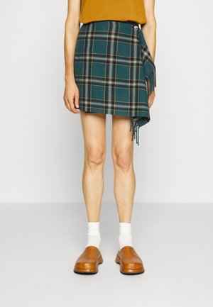 Persoon draagt een teal geruite mini wikkelrok met zijwaartse franjes, een mosterdkleurige top, witte sokken en bruine instapschoenen, staand tegen een effen achtergrond.