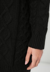 Zwarte gebreide trui met een gestructureerd kabelpatroon, geribde manchetten en een aansluitende pasvorm, die warmte en een gezellig ontwerp benadrukt.