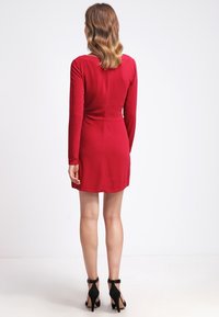 Punainen pitkähihainen mekko, jossa on tiukka siluetti, sileä texture, polvipituinen helma ja pyöreä pääntie. Mustat remmillä varustetut korkokengät.