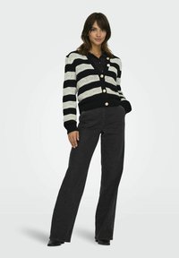 Cardigan rayé noir et blanc avec un motif de tricot texturé, présentant des boutons ronds, associé à un jean foncé taille haute et �à des bottines noires.