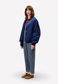 Marine bomberjack met een gladde afwerking, gecombineerd met gestreepte overall en bruine zachte schoenen. Beschikt over brede mouwen en een drukknoopsluiting.