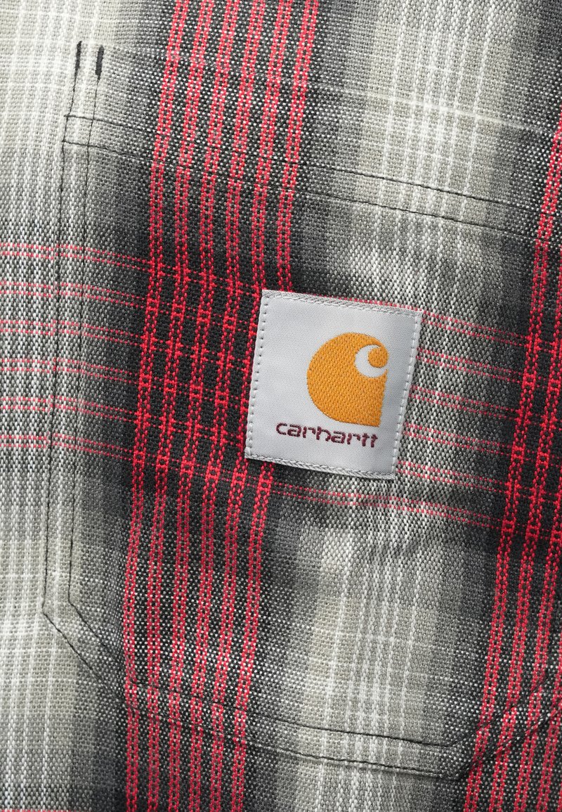 Gray and red plaid fabric featuring a prominent Carhartt patch with an orange logo. The material shows a textured weave and slight sheen.