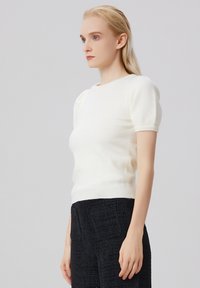 Pull blanc à manches courtes avec un design ajusté et un bord ribbed. Matériau doux avec une texture lisse, porté avec un pantalon noir à taille haute texturé.