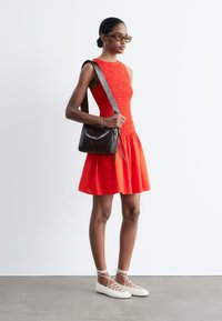Femme portant une robe rouge sans manches, des lunettes de soleil teintées orange, des ballerines blanches à lacets et un sac à bandoulière en cuir noir.