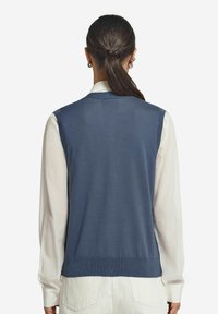 Marinblå ärmlös tröja med slät textur, ribbad fåll och avslappnad passform, bärs över en vit långärmad skjorta.