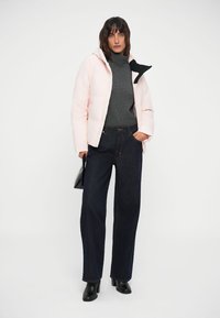 Light pink puffer jacket with a black collar, gray turtleneck, and dark blue straight-leg jeans. Model holds a black handbag.