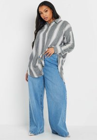 Camisa de botones a rayas gris y blanca con mangas largas, combinada con vaqueros de pierna ancha en azul claro. La camisa tiene un corte relajado y cuello.