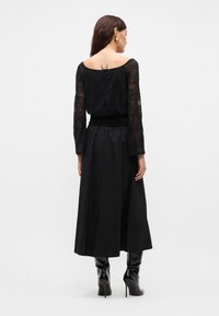 Μαύρη δαντελένια μπλούζα με μακριά μανίκια και στρογγυλή λαιμόκοψη, συνδυασμένη με μία μαύρη σατέν μακριά φούστα. Συνδυασμένη με μαύρες μποτίνες με τακούνι μέχρι το γόνατο.