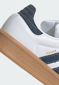 Fehér sportcipő kék csíkokkal, bőr és textil felsőrész, gumitalp, és diszkrét márkázás az oldalon. A talp texturált mintázattal rendelkezik.