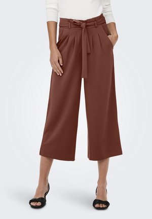 Vrouw draagt high-waist wijde, ingekorte bruine broek met ceintuur, gecombineerd met een witte top en zwarte sandalen met open tenen.