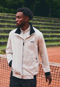 Hellbeige Jacke mit schwarzem Kragen und Reißverschlüssen, auf der Brust ein Logo. Glatte Textur und athletische Passform, kombiniert mit dunklen Hosen.