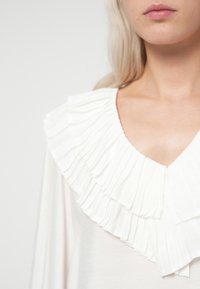 Hvid top med V-hals og en bred, bølget krave. Stoffet ser blødt ud, har en glat struktur og lagdelte plisseringer for ekstra detalje.